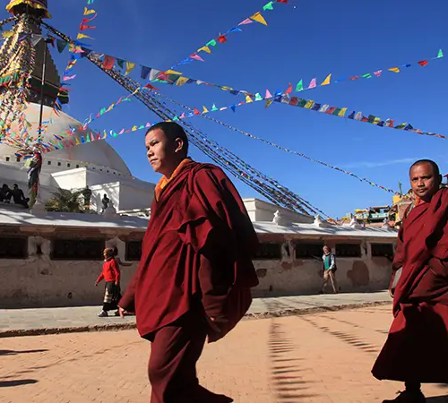 Two Buddhist monks in maroon robes walk past Boudhanath Stupa, with colorful prayer flags overhead under a clear blue sky.