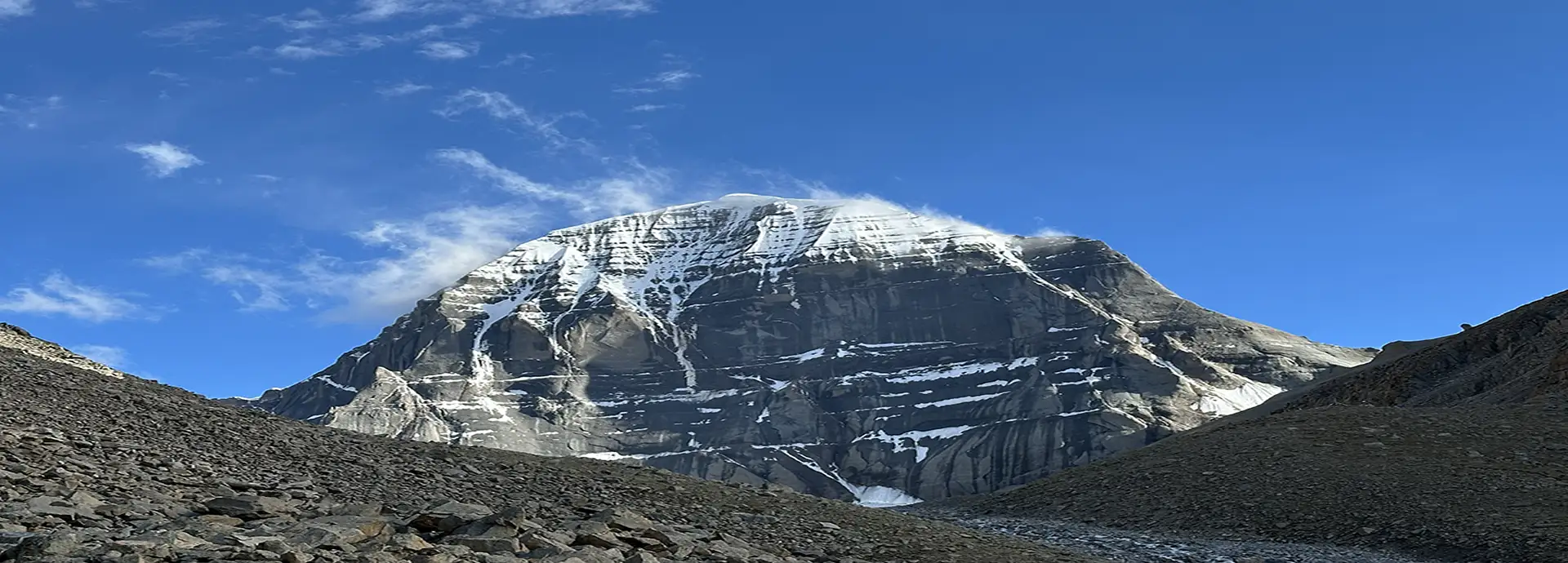 Mount Kailash rising above a rocky high-altitude landscape, with snow-lined ridges under a clear blue sky.