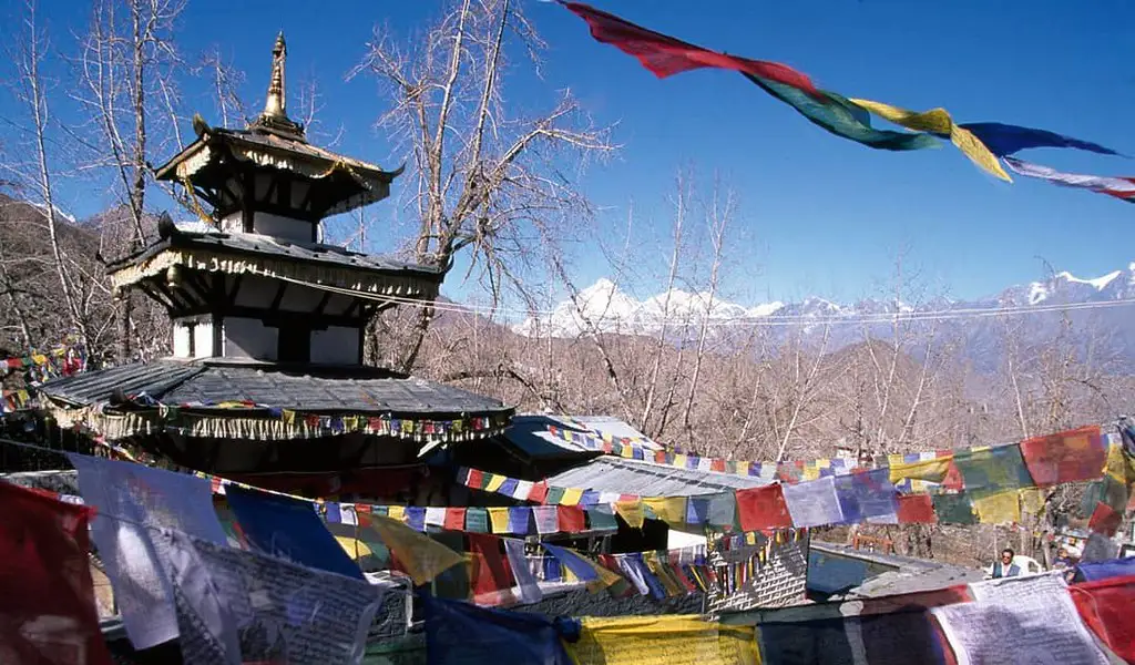 Muktinath Temple in Mustang, Nepal, surrounded by prayer flags with Himalayan mountains visible in the background under a clear blue sky.