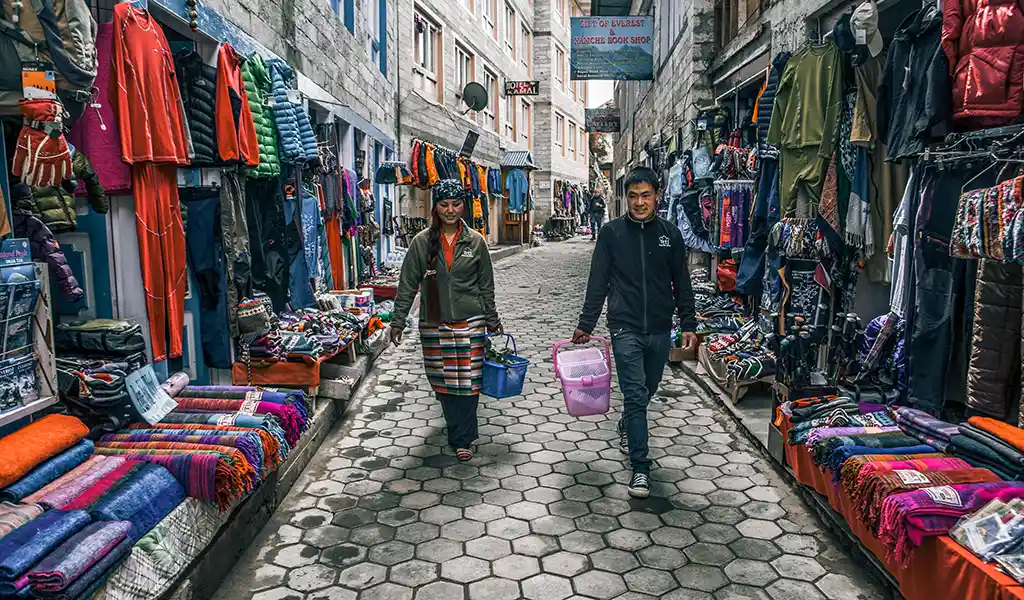 People walking through a narrow street in Namche Bazaar lined with shops selling trekking gear, clothing, and local textiles.