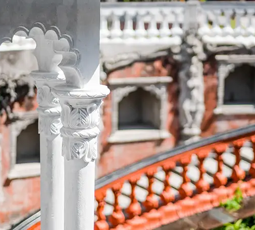 Close-up of a white carved pillar and railing with ornate details, with a red-brick historic building in the background.