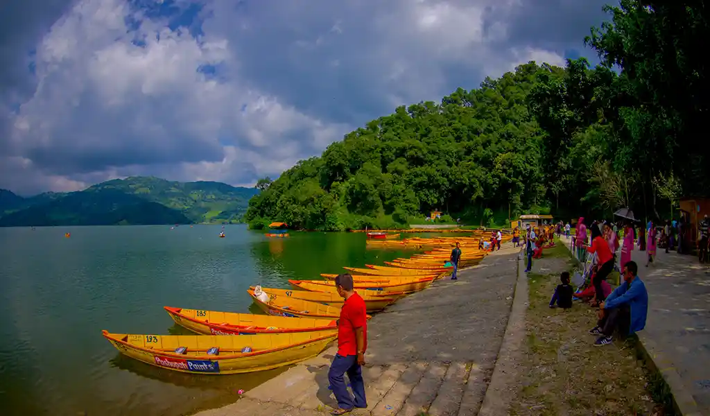 Rowboats are lined up along the shore of Phewa Lake in Pokhara, with visitors on the lakeside path, forested hills, and clouds in the sky.