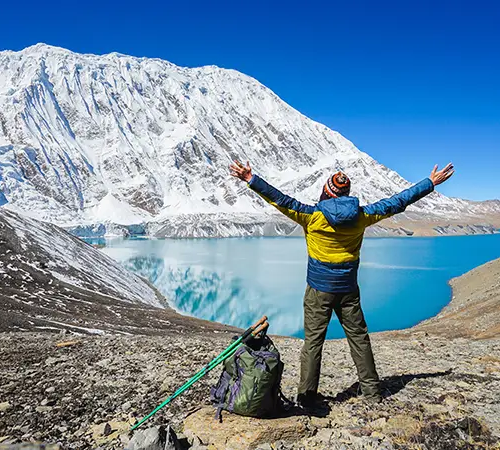 A trekker stands with arms raised above a bright turquoise alpine lake, with a massive snow-covered mountain under a clear blue sky.