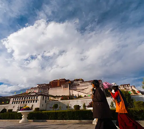 Pilgrims walk along a stone pathway with the Potala Palace rising above them under a dramatic cloudy sky.
