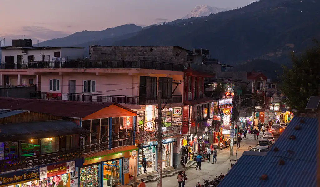 Evening view of Lakeside Street in Pokhara, Nepal, with shops, lights, pedestrians, and mountains in the background.