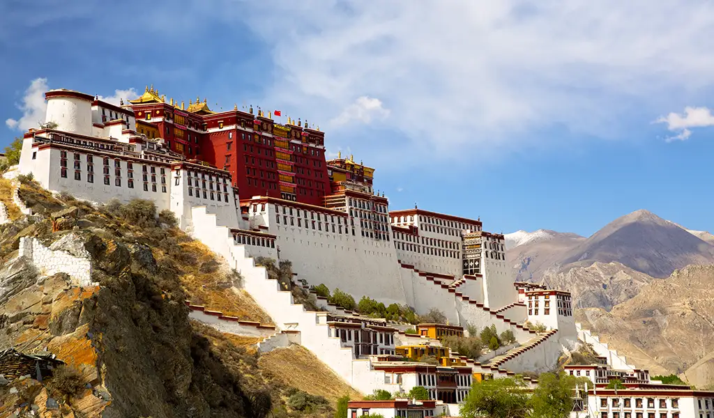 The Potala Palace in Lhasa, Tibet, rises above the hillside with white walls, red palace buildings, and distant mountains under a blue sky.