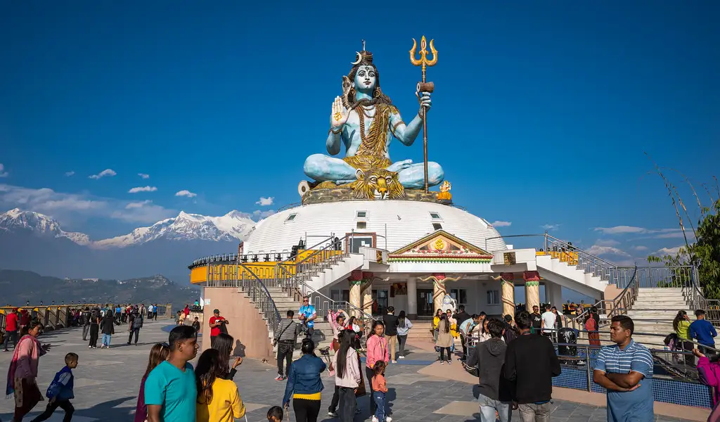 Visitors gather at Pumdikot viewpoint in Pokhara beneath a large statue of Lord Shiva seated on a white dome, with the Annapurna range visible in the background.