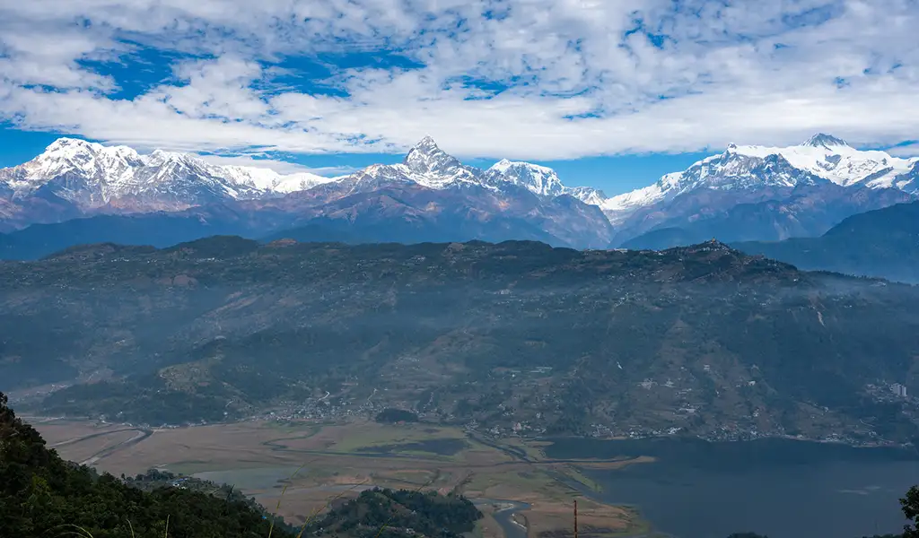 A wide panoramic view from Pumdikot overlooks the Pokhara Valley with Phewa Lake and the snow-capped Annapurna range under a partly cloudy sky.