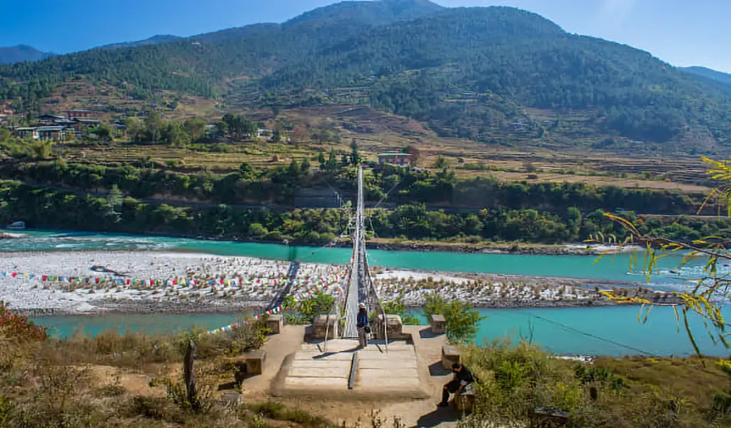 Punakha Suspension Bridge stretching across a turquoise river in Bhutan, with prayer flags and terraced hills in the background.