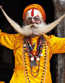 A Hindu sadhu in bright orange clothing and turban poses at a temple doorway, holding up his long curled moustache and wearing layered bead necklaces.