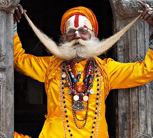 A Hindu sadhu in bright orange clothing and turban poses at a temple doorway, holding up his long curled moustache and wearing layered bead necklaces.