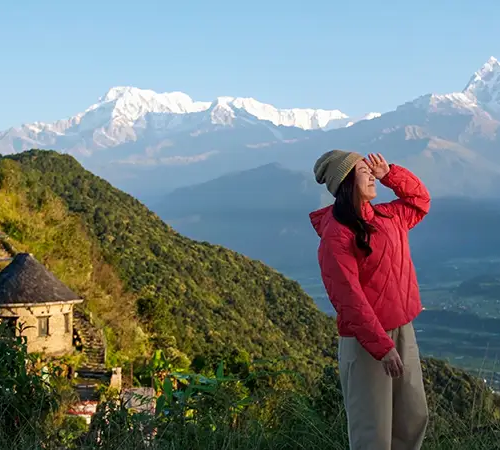 A woman in a red jacket stands on a hillside viewpoint, shading her eyes as she looks toward snow-capped Himalayan mountains.