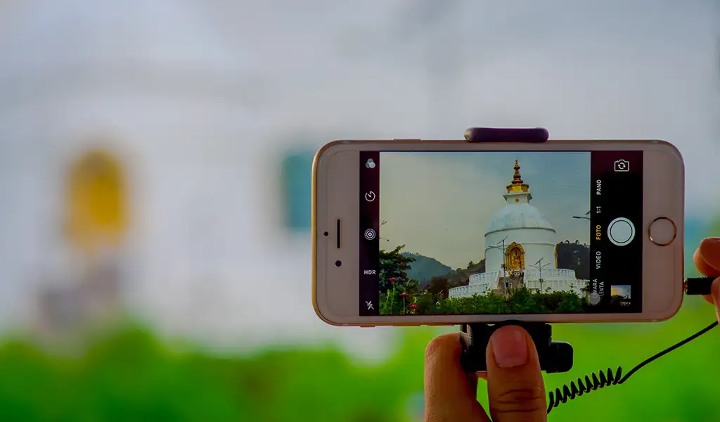 A person photographing the Shanti Stupa (World Peace Pagoda) in Pokhara, Nepal, using a smartphone with the stupa visible on the screen.