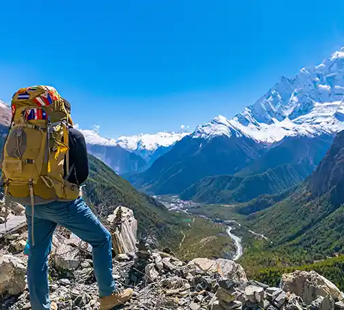 Solo trekker with a backpack standing on a rocky trail, overlooking a deep valley and snow-capped Himalayan peaks along the Annapurna Circuit.