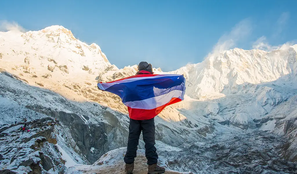 A trekker stands on a snowy ridge holding a flag, facing towering Himalayan peaks under a clear blue sky.