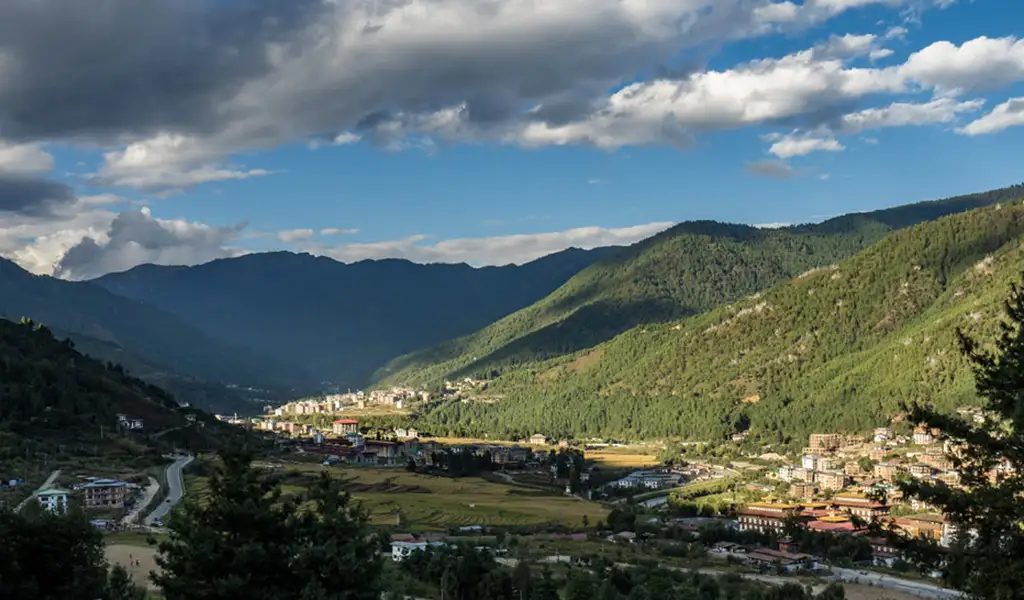 Panoramic view of Thimphu Valley with scattered buildings, terraced fields, and forested hills under dramatic clouds as seen in the Thimphu travel guide