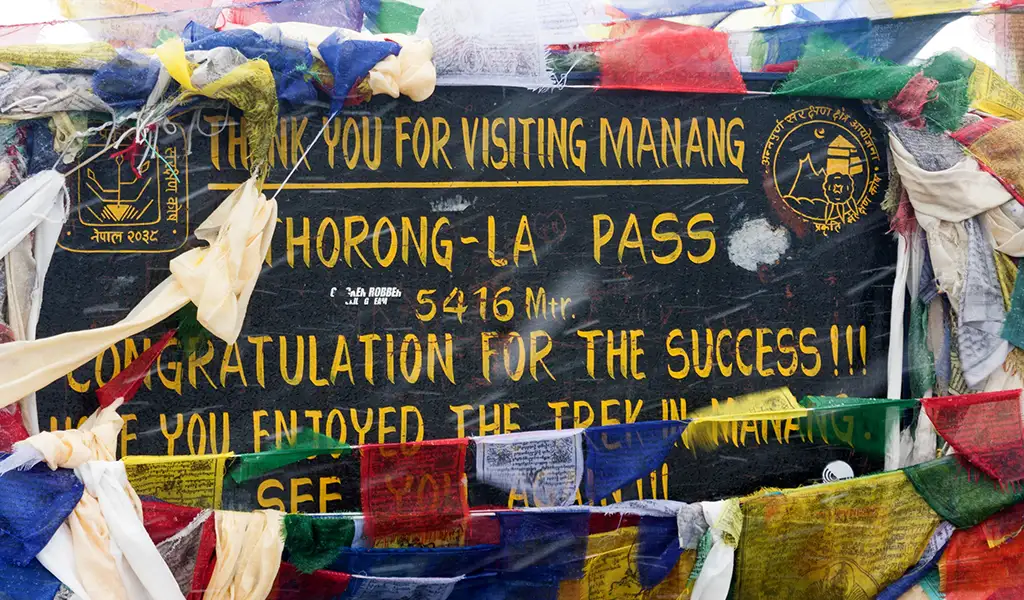 A sign at Thorong La Pass in Manang reads “Thank you for visiting Manang” and “Thorong-La Pass 5416 m,” surrounded by colorful prayer flags.