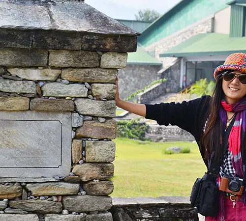 A woman wearing a colorful hat and scarf stands beside a stone monument in a museum courtyard.