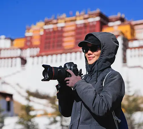 A tourist wearing a jacket and sunglasses holds a camera while standing in front of the Potala Palace.
