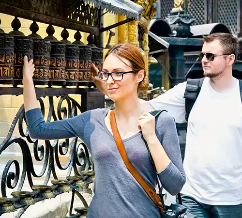 A woman touches a row of prayer wheels while a man walks behind her at a Buddhist temple complex.