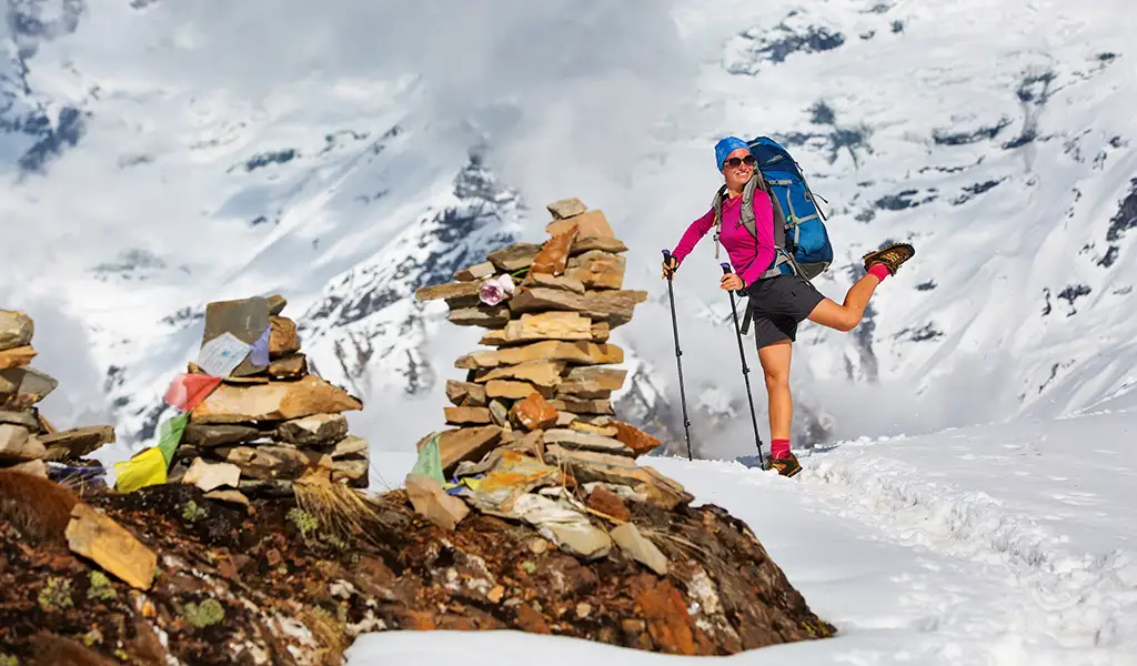 A female trekker poses on a snowy mountain pass while taking Annapurna Circuit trek photos beside stone cairns, prayer flags, and towering peaks in the background.