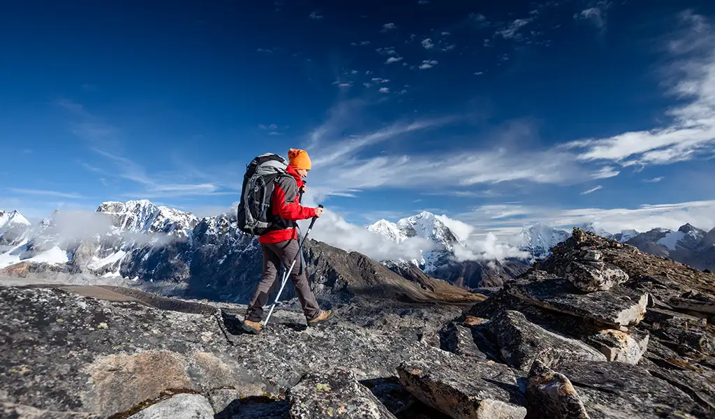A trekker walking across a rocky ridge with a backpack and trekking poles, overlooking snow-covered Himalayan peaks along the Everest Base Camp route.