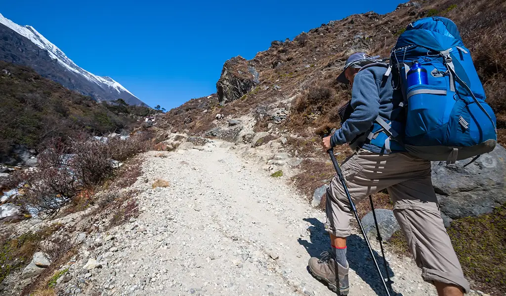 Trekker hiking uphill with a large backpack and trekking poles on a rocky mountain trail, carrying essentials from an Everest Base Camp Trek packing List.