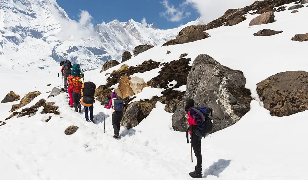 A group of trekkers walks along a snowy trail on the Annapurna Circuit, with rocky slopes and snow-covered Himalayan peaks in the background.