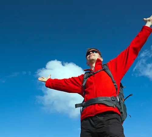 A hiker wearing a red jacket and backpack stands outdoors with arms outstretched, facing the sky under a bright blue sky with clouds.