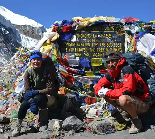 Trekkers resting beside the Thorong La Pass sign at 5,416 meters, surrounded by colorful prayer flags and rocky high-altitude terrain.