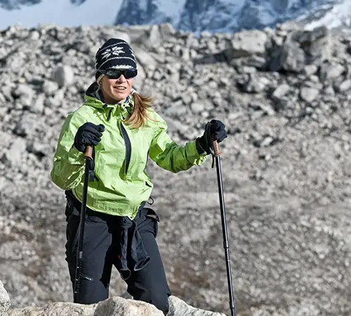 A trekker wearing layered clothing and sunglasses walks across rocky high-altitude terrain using trekking poles in the Everest region.