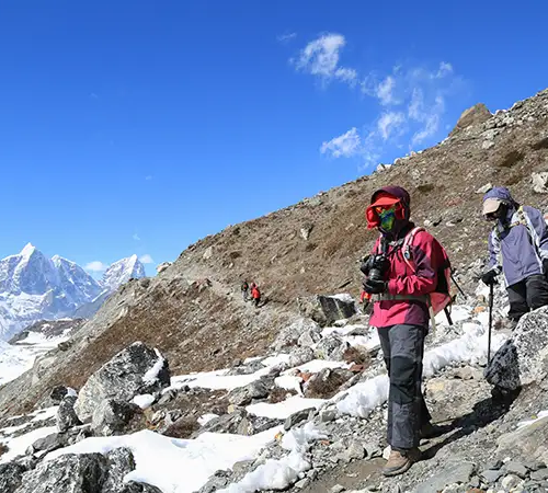 Trekkers wearing full cold-weather gear walk along a rocky, snow-dusted trail in the Everest region, with high Himalayan peaks visible in the distance.