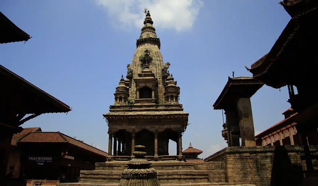 A stone Hindu temple with a tall shikhara rises in the center of a historic square under a clear blue sky.