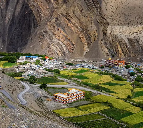 A village in a wide mountain valley with green and yellow fields, winding roads, and steep rocky cliffs in the background.