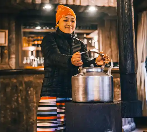 A local woman in Monjo prepares hot tea on a traditional stove inside a teahouse, wearing warm clothing suitable for the mountain climate.