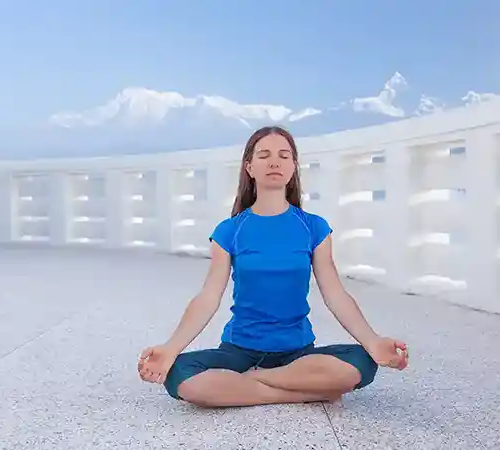 A woman sits cross-legged with eyes closed, meditating on a white terrace with mountains visible in the background.