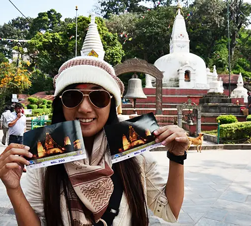 Woman tourist in Nepal holding entrance tickets at a temple complex, smiling in front of white stupas and greenery during sightseeing.