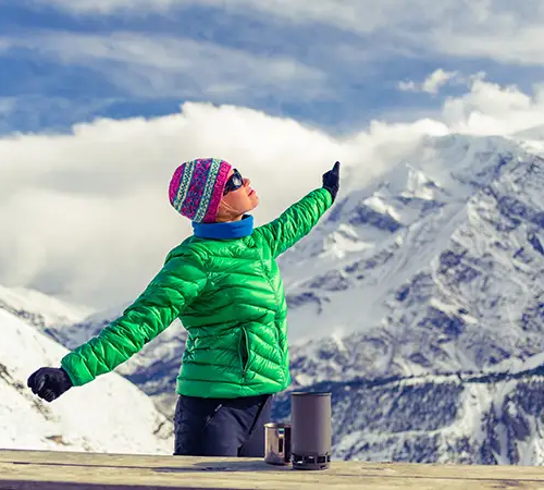 A woman in a green jacket stands with arms outstretched, pointing toward snow-covered mountains at a high-altitude viewpoint.