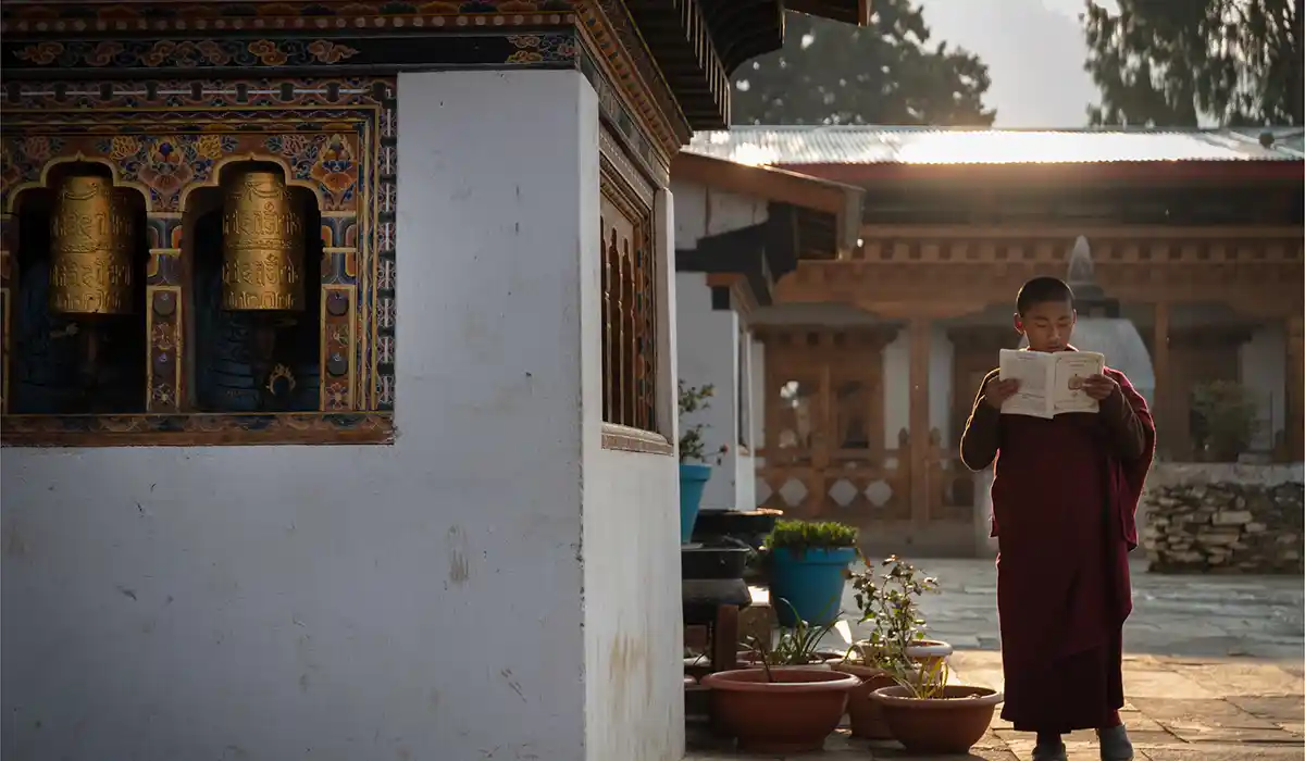 Young Buddhist monk reading scripture near prayer wheels at a monastery courtyard, reflecting Bhutan's Culture and Traditions in daily life.