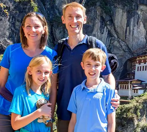 A family of four posing happily on a mountain trail with the Tiger’s Nest Monastery in the background.