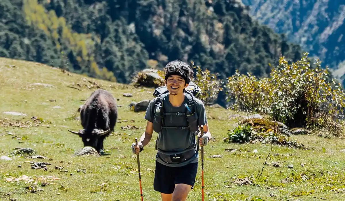 Smiling trekker hiking through alpine meadow during Bhutan Off-Season Travel, with grazing yak and forested Himalayan slopes behind.