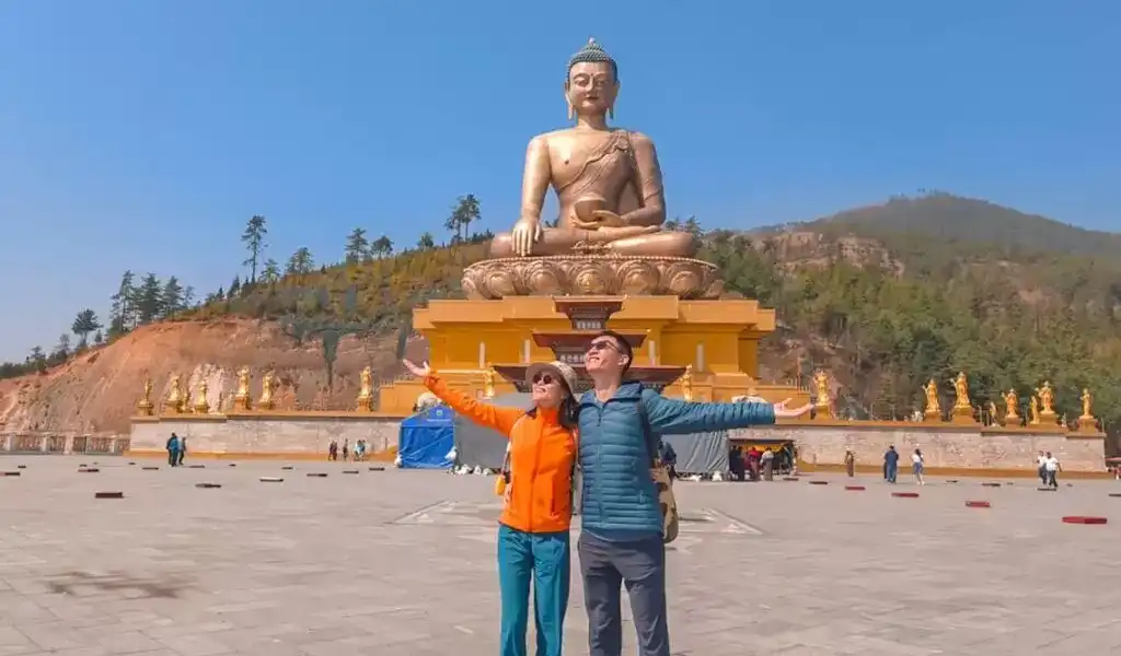 A happy couple standing before the massive golden Buddha Dordenma statue on a romantic Bhutan honeymoon tour.