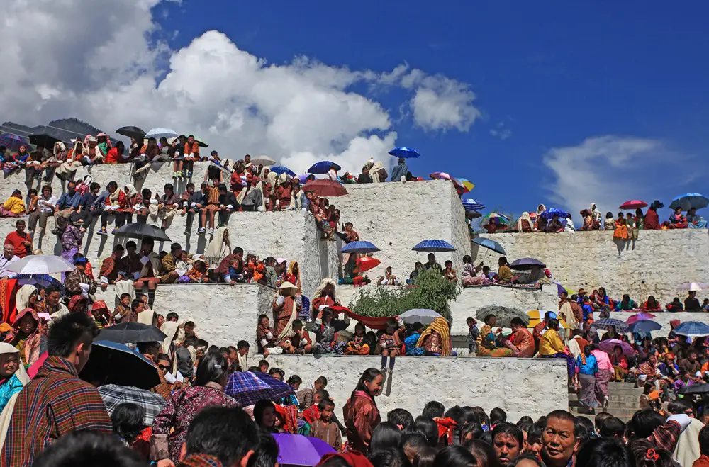 Crowds gathered on white terraces during Thimphu Tsechu Festival in Bhutan under a clear blue sky.