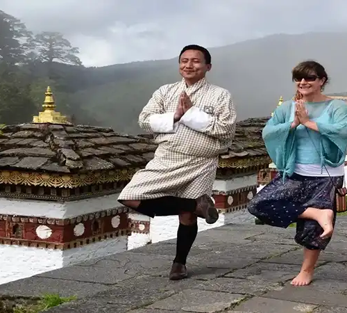A man and woman practice a yoga tree pose near mountain stupas on a Bhutan spiritual tour.