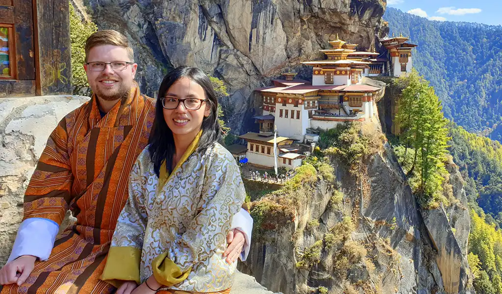A couple in traditional Kira and Gho outfits takes a photo with Tiger’s Nest Monastery on a Bhutan honeymoon tour.