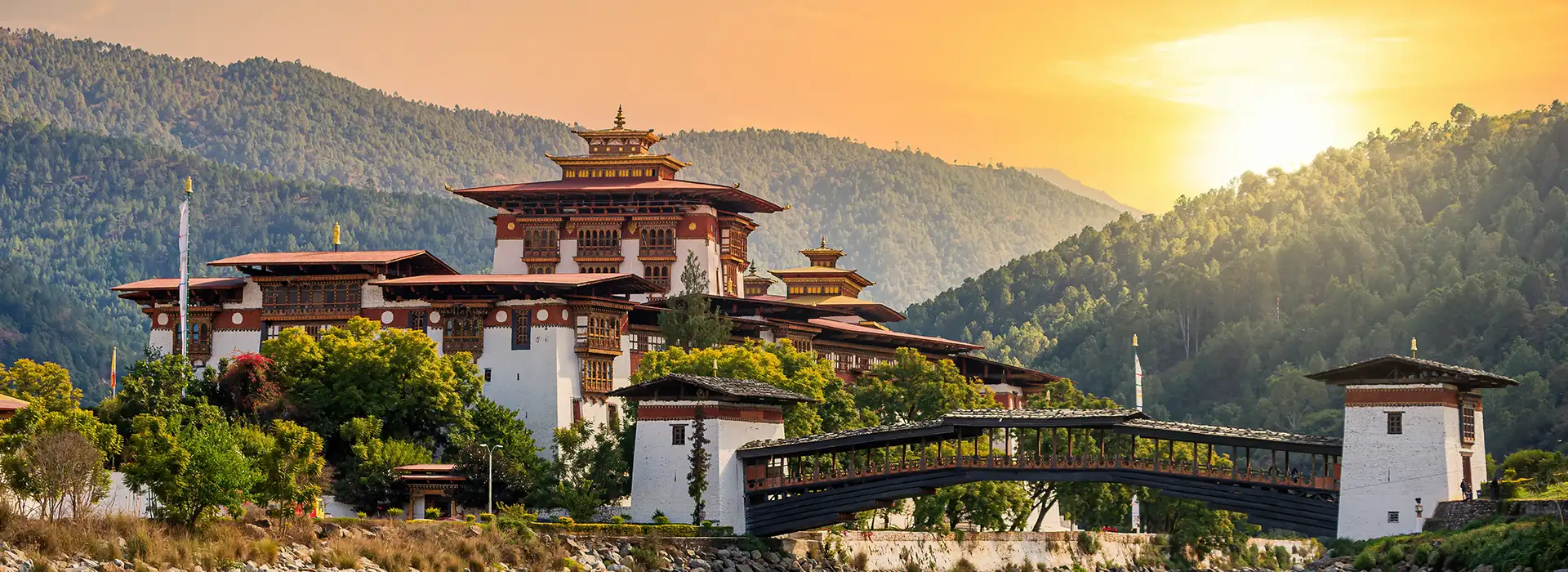 A wide view of the historic Punakha Dzong fortress in Bhutan at sunset, situated by a river with a traditional wooden cantilever bridge, preserved through the Sustainable Development Fee (SDF).