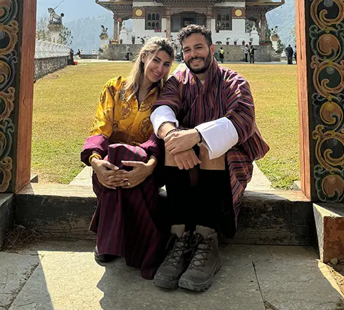 Couple wearing traditional Bhutanese dress seated in a monastery courtyard, experiencing local culture featured in a Bhutan accommodation guide.