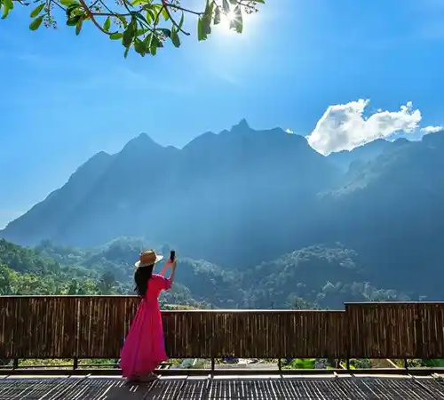 Woman in pink dress photographing Himalayan peaks during Bhutan Off-Season Travel from scenic wooden viewpoint under clear blue sky.