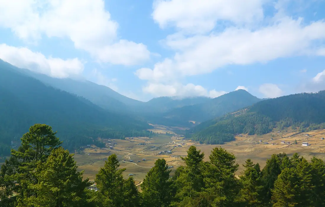 Panoramic view of Phobjikha Valley with pine forests, open farmland, scattered traditional houses, and misty Himalayan hills under a blue sky in Bhutan.