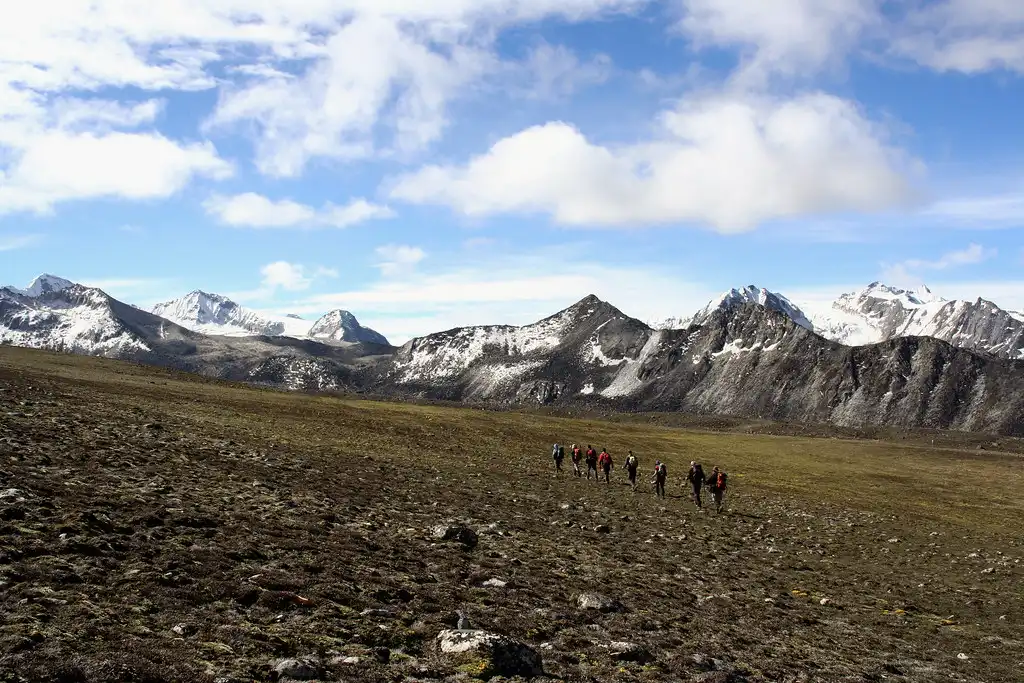 A line of trekkers walking across a vast, rocky high-altitude plateau with snow-dusted Himalayan peaks in the background during a Druk path trek.